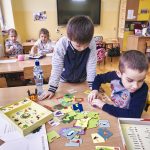 Dwóch chłopców w kolorowych swetrach gra w puzzle rozłożone na stole. Z tyłu dwie dziewczynki siedzą przy stole