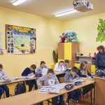 Nauczycielka z książką w ręce patrzy na dzieci siedzące w ławkach i odrabiające zadanie