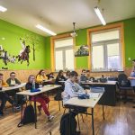 Dzieci siedzą w ławkach w klasie. Na ścianie koloru zielonego namalowana scena bitewna