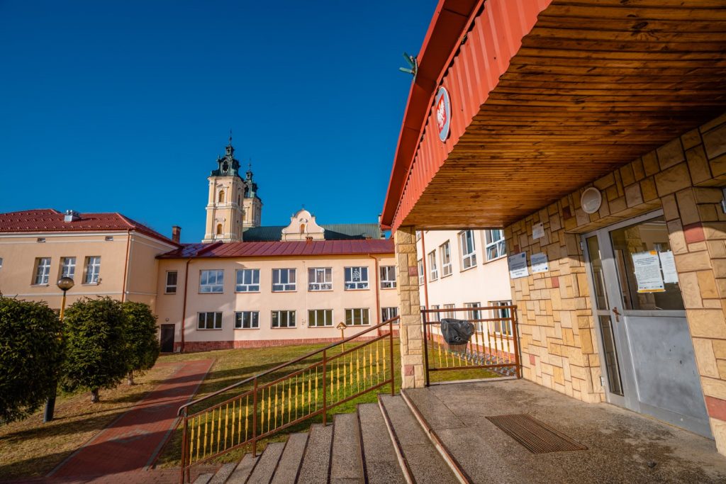 Wejście do budynku szkolnego. W głębi inne budunki i barokowy kościół z dwoma wieżami