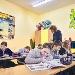 Dzieci siedzą w ławkach i pisza w zeszytach. Z tyłu na ścianie wisi mapa Polski